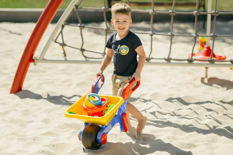 A small boy pushing a toy wheelbarrow in a sandy playground